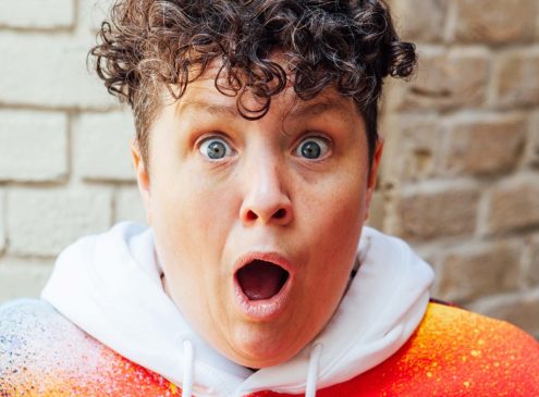 Jess Thom, a white woman with short curly brown hair wearing a multi-coloured hoodie, with a surprised expression on her face.