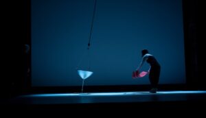 A swinging funnel deposits sand on a sheet on a stage as a performer does something undetermined with a large piece of red cardstock. A second performer stands just at the edge of the frame.