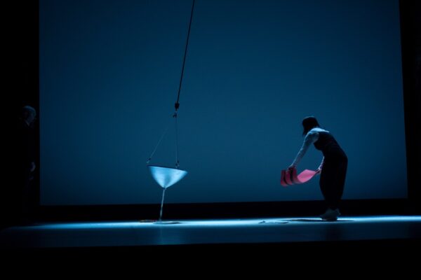 A swinging funnel deposits sand on a sheet on a stage as a performer does something undetermined with a large piece of red cardstock. A second performer stands just at the edge of the frame.