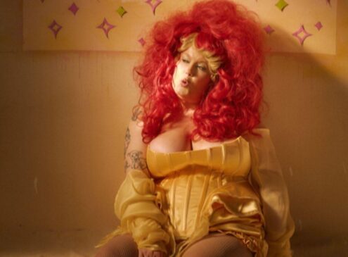 A woman with bright red hair and a yellow dress sits on the floor