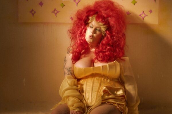 A woman with bright red hair and a yellow dress sits on the floor