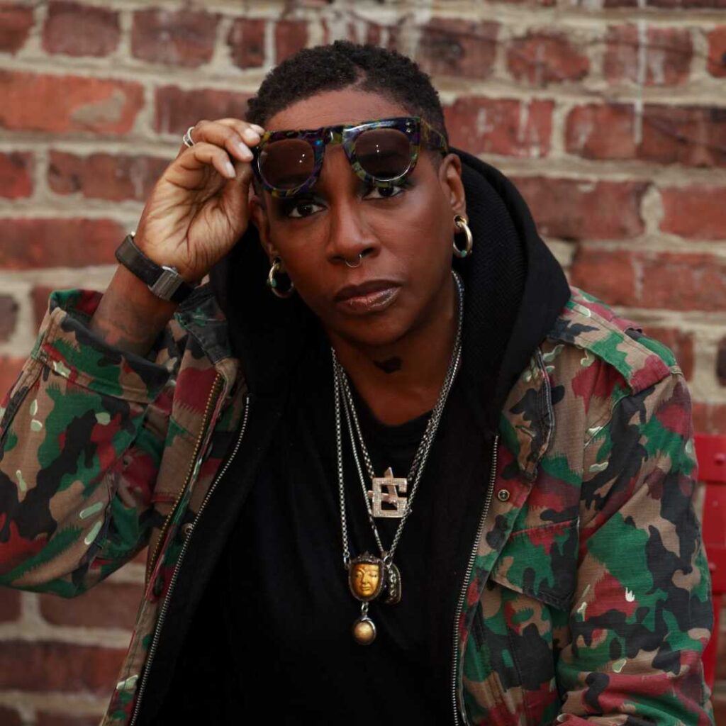 A woman wearing a camo jacket holding her sunglasses looking at the camera