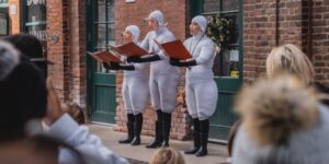 A group of three people dressed as sheep holding books outside and singing in front of a crowd