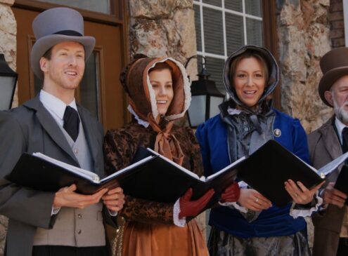 A group of four carolers wearing period attire holding books and singing
