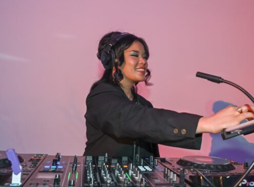 A woman spinning music at a DJ booth against a pink background
