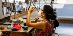 A woman wearing a mask working on a jewelry piece in a studio