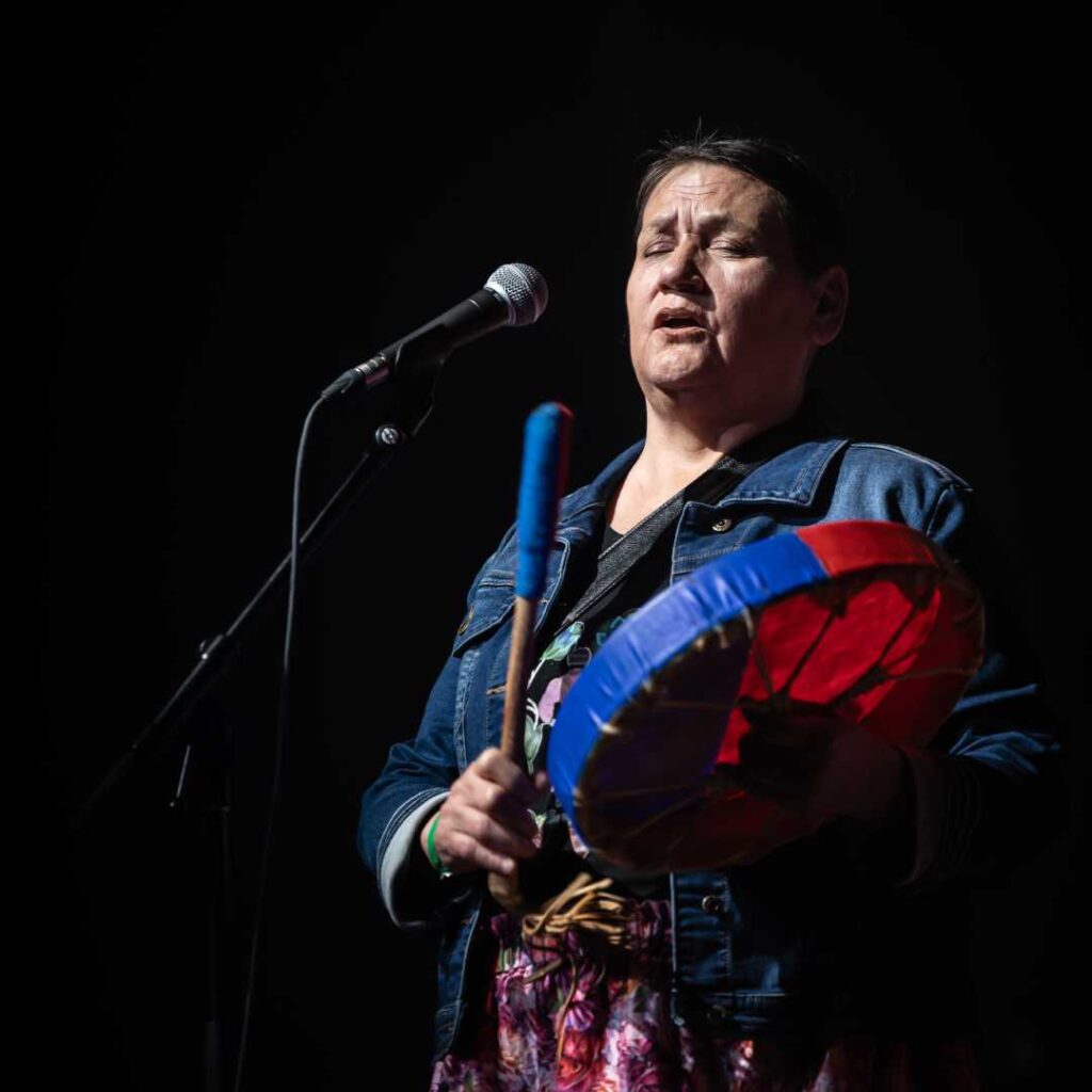 A woman holding a colourful percussion instrument performing on stage against a dark background