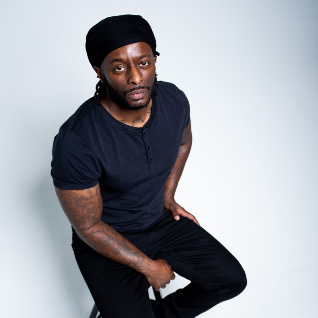 A man wearing a black shirt and hat sitting on a stool posing for the camera