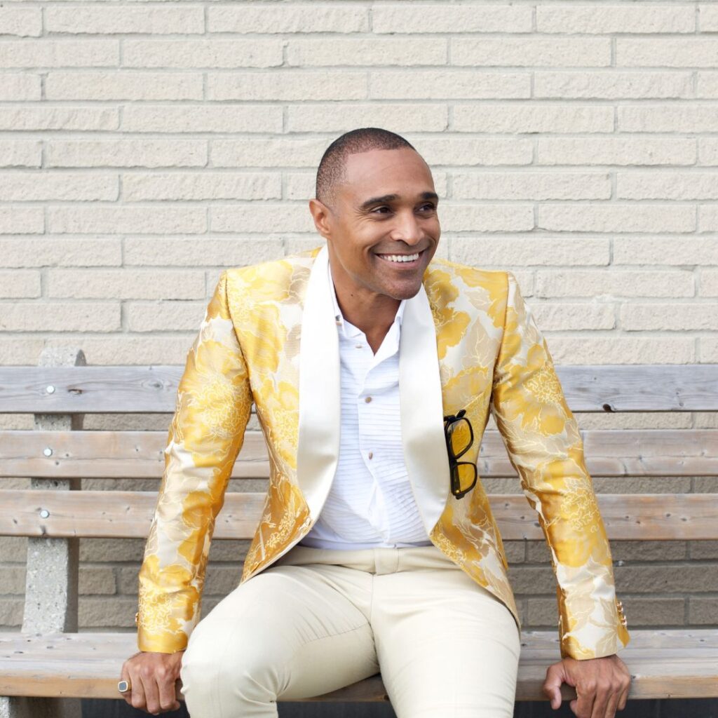 A man sitting on a bench outdoors wearing a yellow blazer smiling