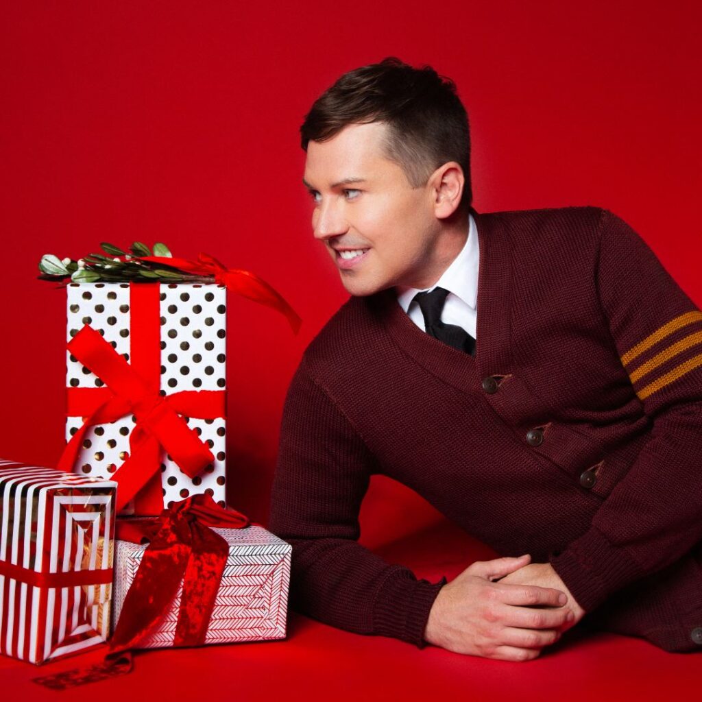 A man wearing a red sweater lying next to wrapped presents against a red background