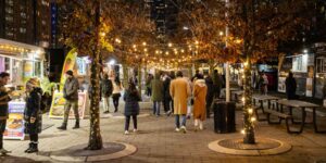 An outdoor food market with crowds walking past food trucks and holiday lights