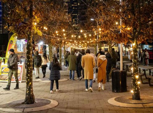 An outdoor food market with crowds walking past food trucks and holiday lights