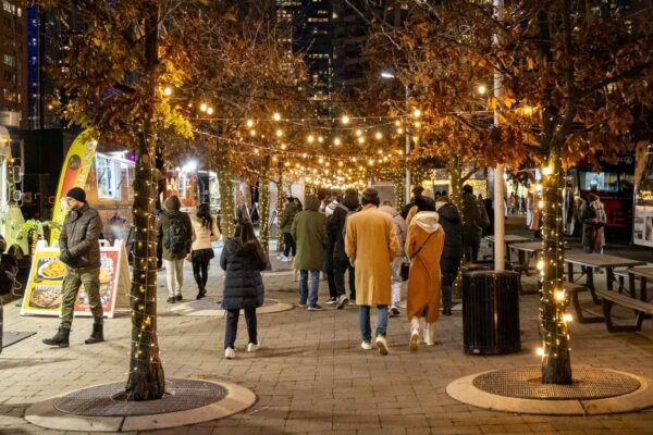 An outdoor food market with crowds walking past food trucks and holiday lights