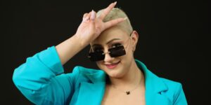 A person wearing a bright blue jacket holding a peace sign against a dark background