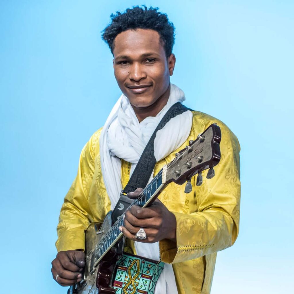 A man holding a musical instrument wearing a yellow jacket against a blue background