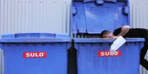 A man wearing black leaning into a bright blue garbage bin