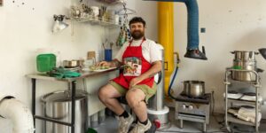 An artist wearing a red apron sitting down in a studio and posing for the camera
