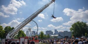 A man suspended by a crane doing a performance before a crowd outdoors.