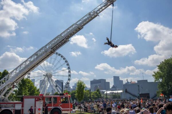 A man suspended by a crane doing a performance before a crowd outdoors.