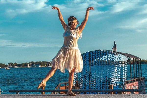 A woman in 1920s attire dancing and posing in front of a waterfront outdoors