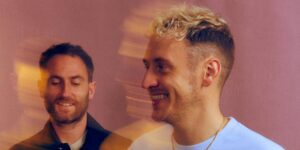 A close-up of two musicians smiling against a pink background.