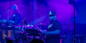 Two artists on stage playing keyboard and drums against a purple light