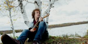 A musician sitting against a tree o utside holding a guitar looking off into the distance.