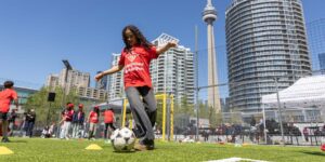 A girl kicking a soccer ball on an outdoor pitch with the CN tower in the background