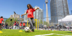 A girl kicking a soccer ball on an outdoor pitch with the CN tower in the background