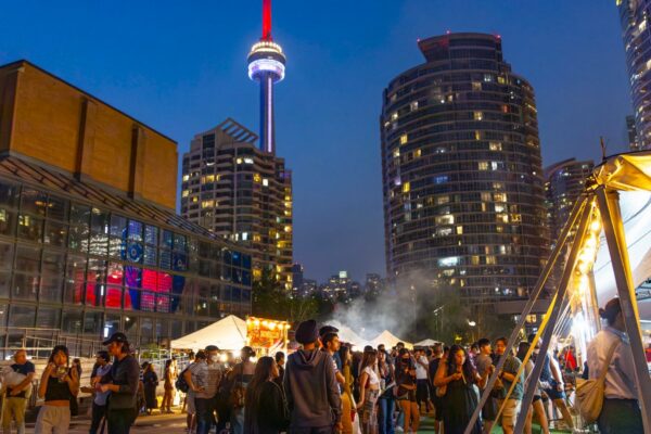 A nighttime shot of a crowd at an outdoor food market with the CN tower in the background